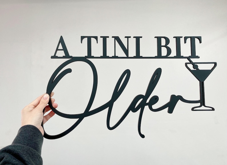 May include: A black metal sign with the text "A TINI BIT Older" in a cursive font. A martini glass with a straw is to the right of the text.