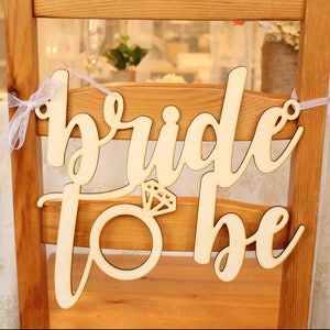 May include: Wooden sign with the words "bride to be" in cursive script. A ring with a diamond is attached to the sign.