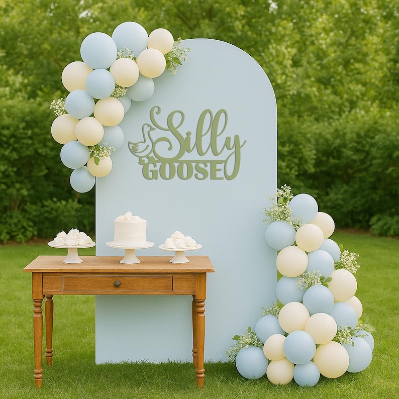 Puede incluir: Un tel&oacute;n de fondo azul claro con las palabras "Silly Goose" en escritura verde, decorado con globos pastel y detalles florales. Una mesa de madera exhibe un pastel blanco y postres, creando una escena festiva.
