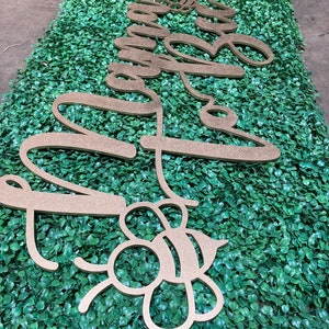 May include: Gold-colored wooden cutouts spelling "Sweet Bee" in cursive script and a bee-shaped design. The cutouts are placed on a green, leafy background, likely for decorative purposes.