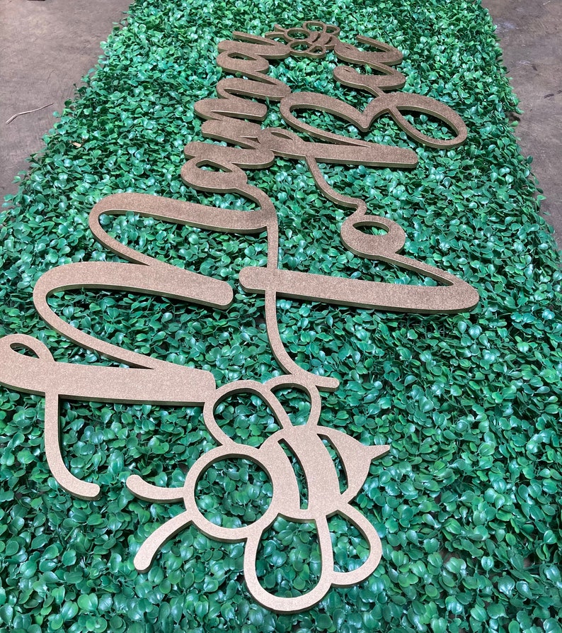 May include: Gold-colored wooden cutouts spelling "Sweet Bee" in cursive script and a bee-shaped design. The cutouts are placed on a green, leafy background, likely for decorative purposes.