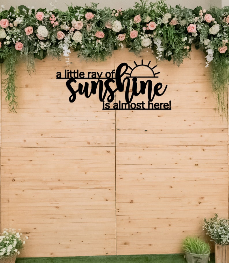 May include: A black wooden sign with the text "a little ray of sunshine is almost here!" with a sun graphic. The sign is hanging on a light brown wooden wall with a floral garland above it.