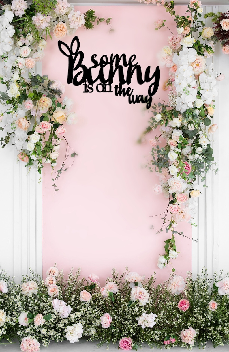 May include: A pink background with a black sign that reads "Some bunny is on the way". The sign is surrounded by white and pink flowers.