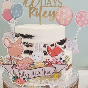 May include: A two-tiered cake with white frosting and a wooden topper that reads "Happy 100 Days Riley." The cake is decorated with animal cutouts, balloons, and a banner with the names "Riley Aria Han." The cake sits on a decorative gold stand.