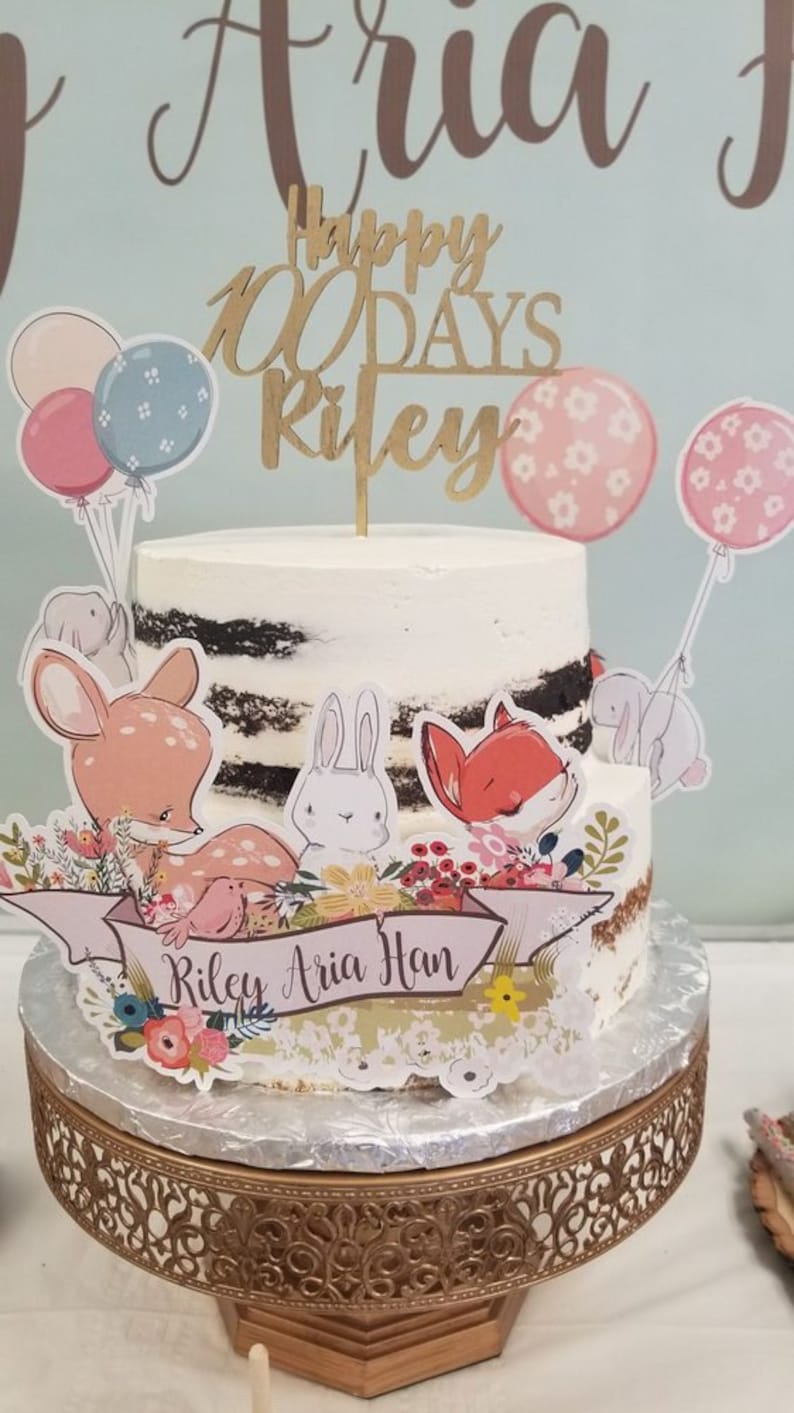 May include: A two-tiered cake with white frosting and a wooden topper that reads "Happy 100 Days Riley." The cake is decorated with animal cutouts, balloons, and a banner with the names "Riley Aria Han." The cake sits on a decorative gold stand.