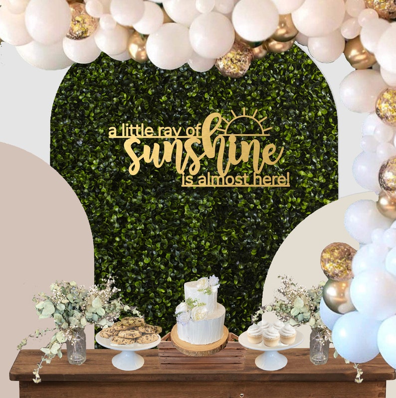 May include: A baby shower dessert table with a green backdrop and gold balloons. The table has a wooden surface and is decorated with cookies, cupcakes, and a two-tiered cake. A gold sign reads "a little ray of sunshine is almost here!"