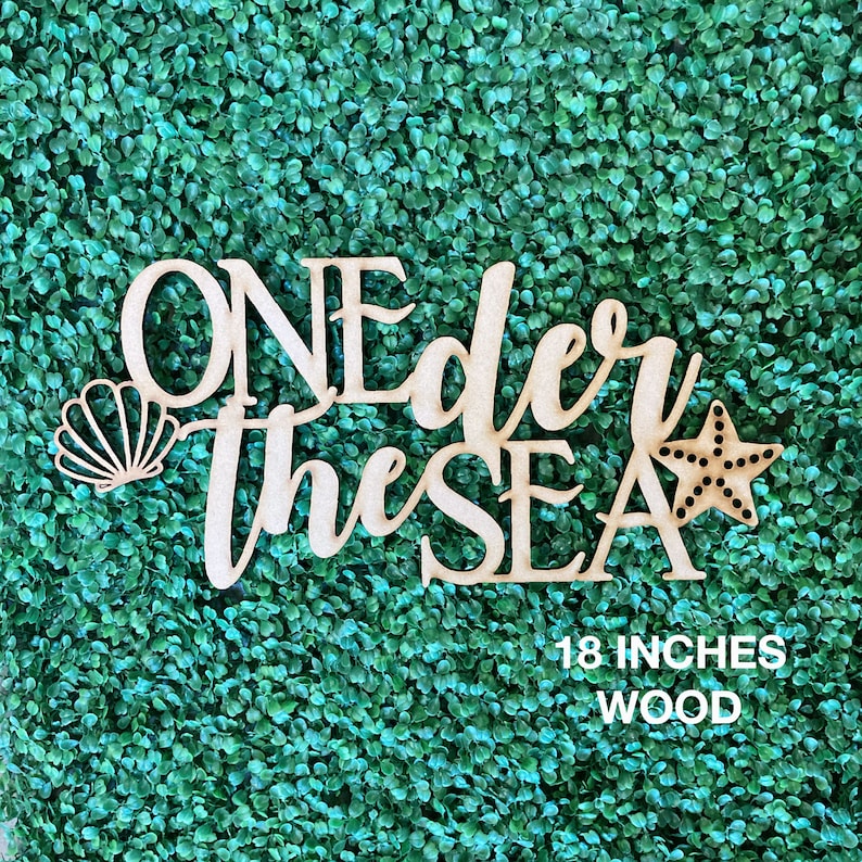 May include: Wooden sign with the text "One der the SEA" in a cursive font. The sign is decorated with a seashell and a starfish. The sign is 18 inches tall and made of wood.