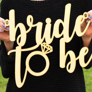May include: Wooden sign with the text "bride to be" in cursive script. A diamond ring is depicted in the center of the sign.