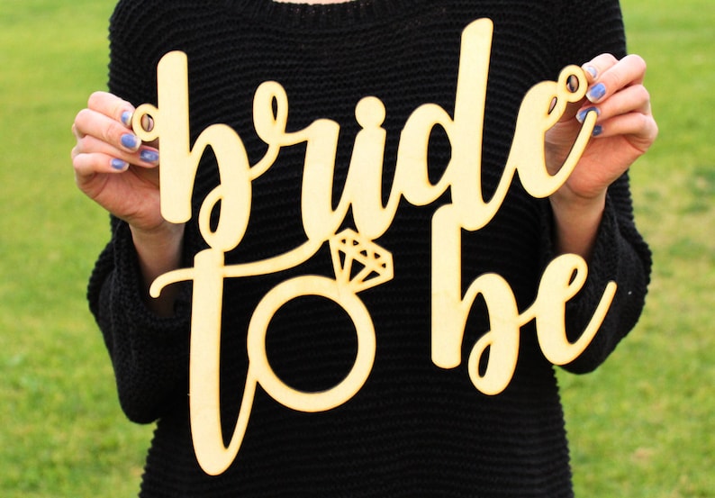 May include: Wooden sign with the text "bride to be" in cursive script. A diamond ring is depicted in the center of the sign.