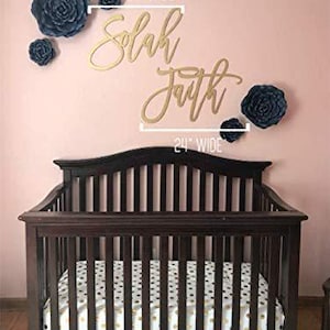 May include: A dark brown wooden crib with a mattress featuring a white and gold pattern. Above the crib, on a pink wall, are gold script words that read "Solah Faith" and several dark blue paper flowers.