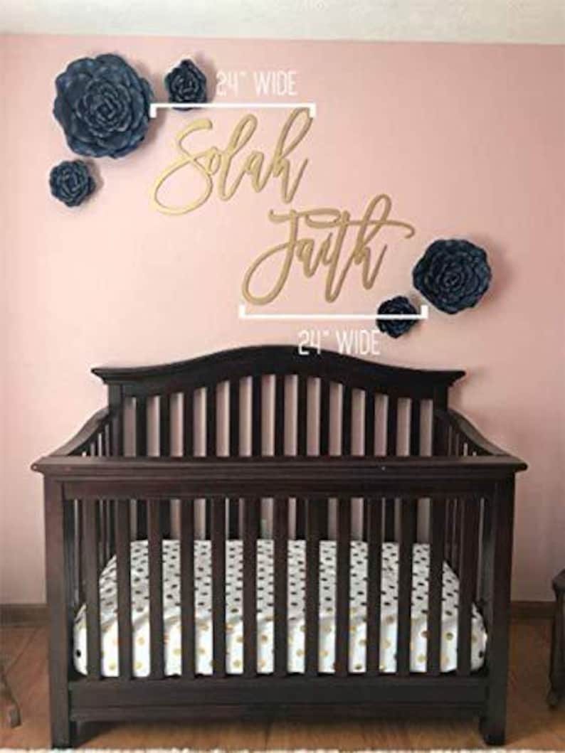 May include: A dark brown wooden crib with a mattress featuring a white and gold pattern. Above the crib, on a pink wall, are gold script words that read "Solah Faith" and several dark blue paper flowers.