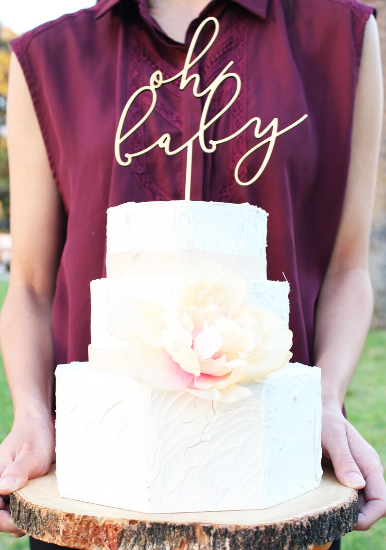 May include: A three-tiered white cake with a large, light pink and cream-colored flower. The cake is topped with a wooden "oh baby" cake topper. The cake is on a wooden serving board.