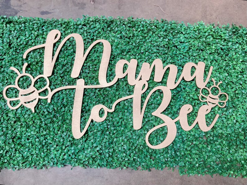 May include: Wooden sign with the words "Mama to Bee" in cursive script, with bee illustrations on either side. The sign is light brown and set against a green textured background.