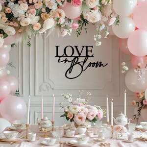 Op de afbeelding: Een zwart houten bord met de tekst "LOVE in BLOOM" in een cursief schrift. Het bord hangt aan een witte muur met een bloemenkrans en roze ballonnen op de achtergrond. Een tafel is gedekt met witte tafelkleden, roze bloemen en theekopjes.
