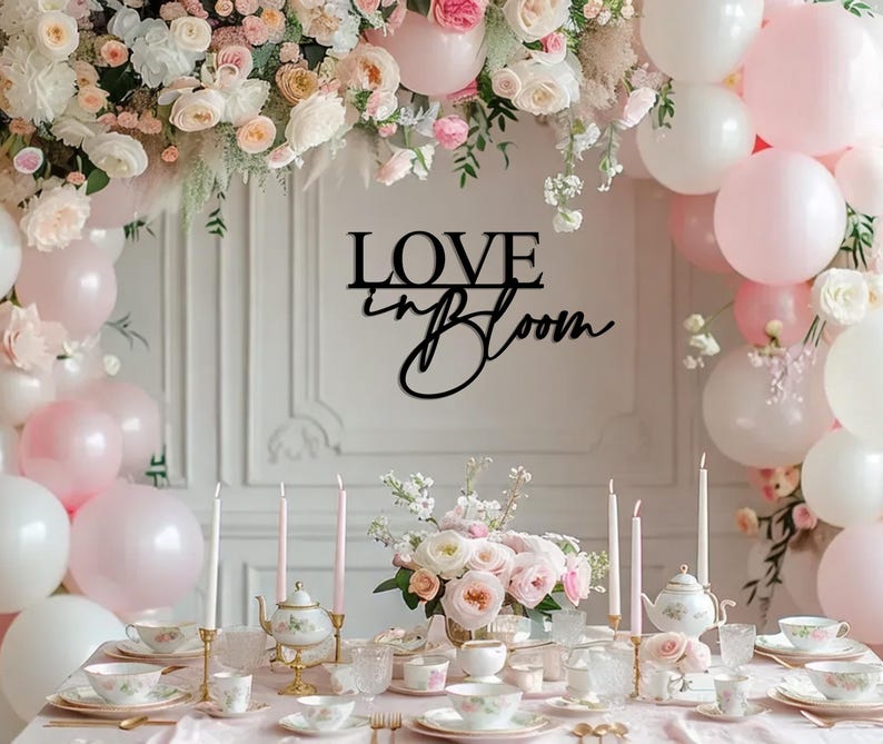 Op de afbeelding: Een zwart houten bord met de tekst "LOVE in BLOOM" in een cursief schrift. Het bord hangt aan een witte muur met een bloemenkrans en roze ballonnen op de achtergrond. Een tafel is gedekt met witte tafelkleden, roze bloemen en theekopjes.