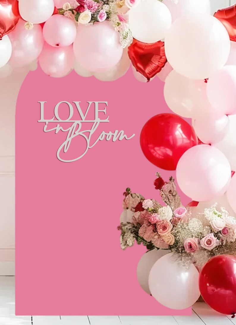 Op de afbeelding: Een roze achtergrond met een wit houten bord waarop "LOVE in BLOOM" staat. De achtergrond is versierd met een krans van roze en rode ballonnen.