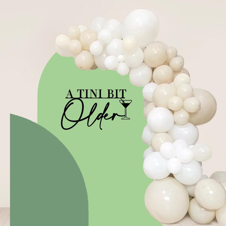 May include: A green and white balloon arch with the text "A TINI BIT Older" and a martini glass icon. The balloons are white and cream.
