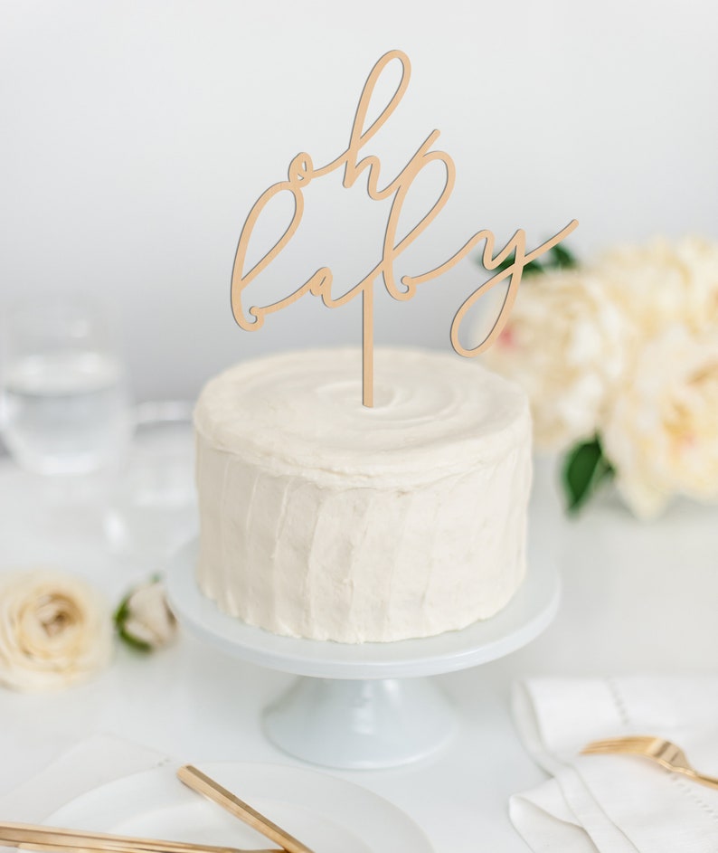 May include: A white frosted cake with a wooden cake topper that reads "oh baby" in cursive script. The cake sits on a white cake stand, with flowers and place settings in the background. A celebratory dessert.
