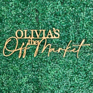 Może przedstawiać: Jasnobrązowy drewniany znak z napisem "OLIVIA'S the Off Market" w połączeniu czcionek blokowych i kursywy. Znak jest umieszczony na tle gęstego, zielonego listowia.