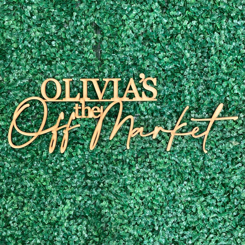 Może przedstawiać: Jasnobrązowy drewniany znak z napisem "OLIVIA'S the Off Market" w połączeniu czcionek blokowych i kursywy. Znak jest umieszczony na tle gęstego, zielonego listowia.