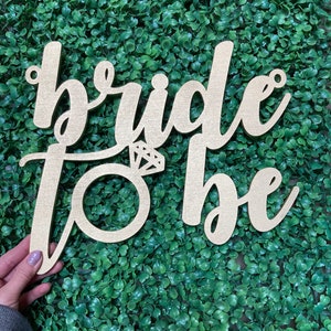 May include: Wooden sign with the words "bride to be" in cursive script. The word "bride" is larger than the word "to be". A ring with a diamond is in the center of the sign.