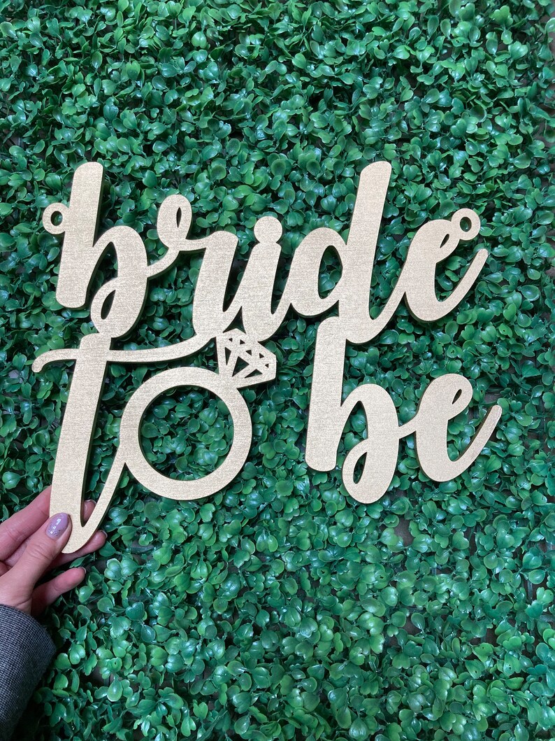 May include: Wooden sign with the words "bride to be" in cursive script. The word "bride" is larger than the word "to be". A ring with a diamond is in the center of the sign.