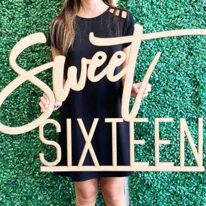 May include: A wooden sign with the words "Sweet Sixteen" in a cursive font. The sign is held by a person in front of a green wall.