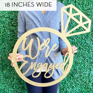 May include: A gold wooden ring with the words "We're Engaged" in cursive script. The ring is topped with a diamond shape. The sign is 18 inches wide.