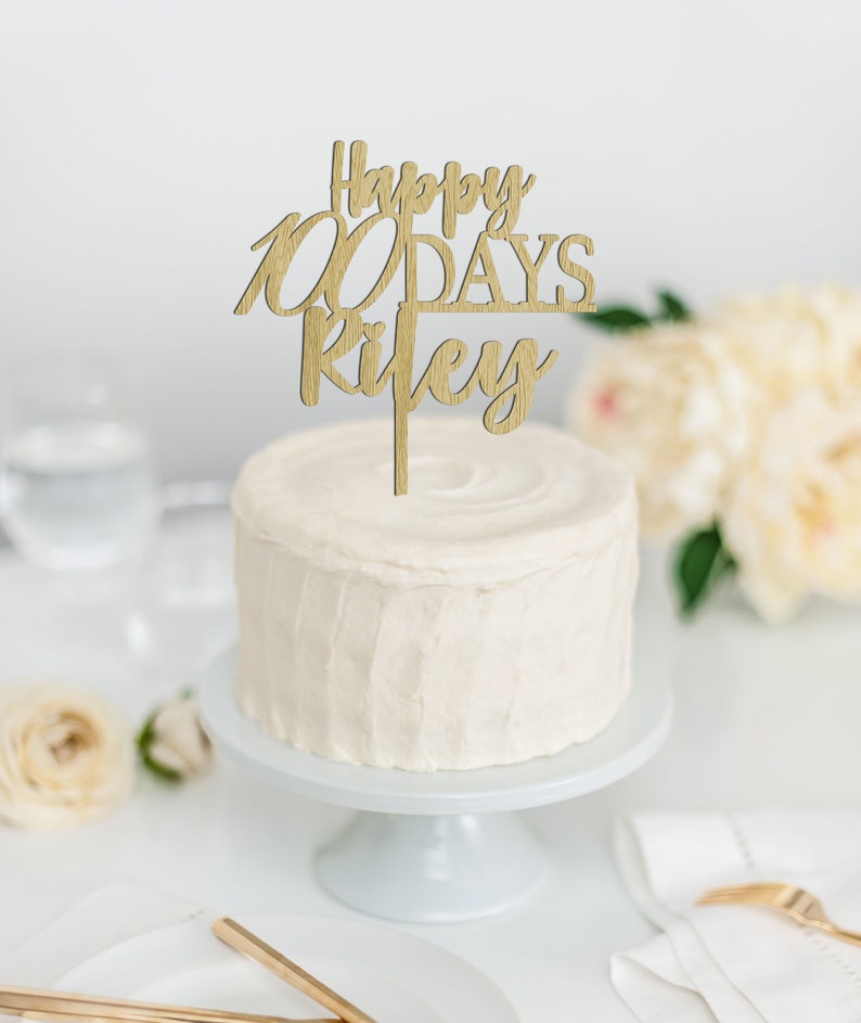May include: A white frosted cake with a wooden cake topper that reads "Happy 100 Days, Ryley". The cake sits on a white cake stand. The background includes flowers, a glass of water, and a white table setting.