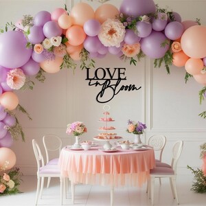 Op de afbeelding: Een tafeldecoratie voor een bruiloft of een bruidsdouche met een roze tafelkleed en een ballonnenboog gemaakt van pastelpaarse en perzikkleurige ballons. De boog is versierd met groen en bloemen. Een zwart houten bord met de tekst "LOVE in BLOOM" hangt boven de tafel.