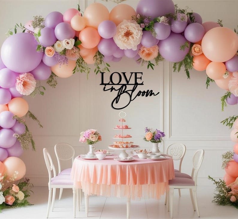 Op de afbeelding: Een tafeldecoratie voor een bruiloft of een bruidsdouche met een roze tafelkleed en een ballonnenboog gemaakt van pastelpaarse en perzikkleurige ballons. De boog is versierd met groen en bloemen. Een zwart houten bord met de tekst "LOVE in BLOOM" hangt boven de tafel.