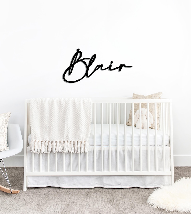 May include: A black wooden sign with the name "Blair" in cursive script is mounted above a white crib. The crib is decorated with a white blanket, a pillow, and a stuffed bunny. A white rocking chair is visible on the left.