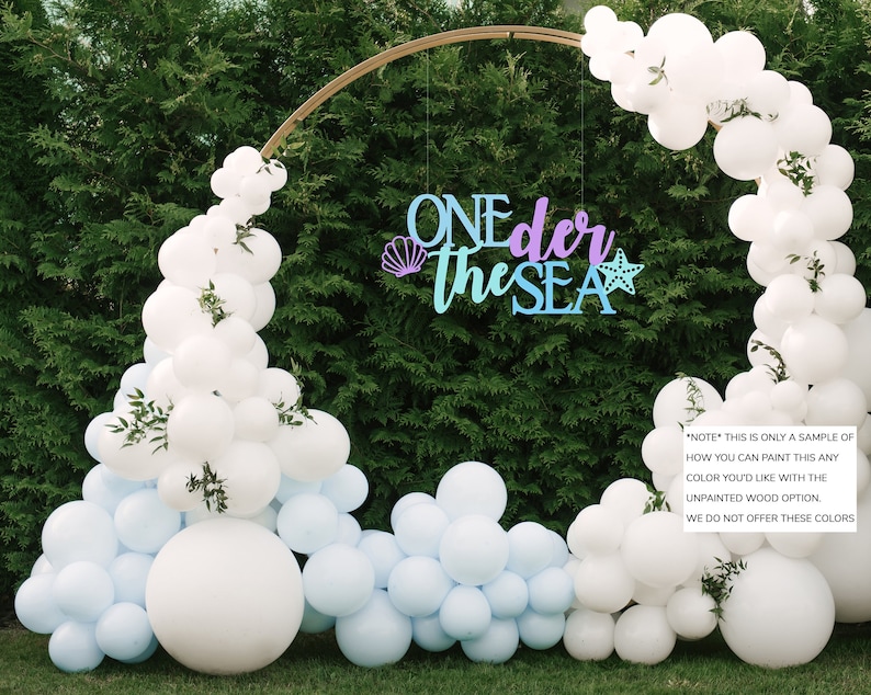May include: A wooden sign with the text "ONEder the SEA" in blue and purple paint. The sign is decorated with a seashell and a starfish. The sign is hanging from a gold archway decorated with white and light blue balloons.