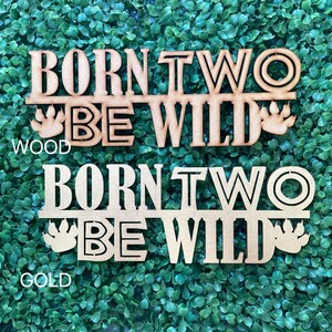 May include: Two wooden signs with the text "Born Two Be Wild" in a bold font. The top sign is natural wood and the bottom sign is gold. Both signs feature a paw print on either side of the text.