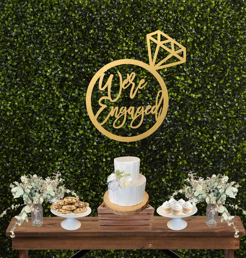 May include: A gold wooden sign with the words "We're Engaged" in a circle with a diamond ring on top. The sign is hanging on a green wall with a white cake and cupcakes on a wooden table in front of it.