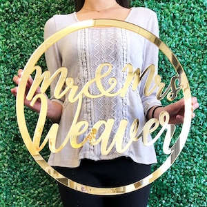 May include: A gold-colored, circular sign with the words "Mr & Mrs Weaver" in elegant script. The sign is held in front of a green hedge background, creating a striking contrast. The sign is made of a reflective material.
