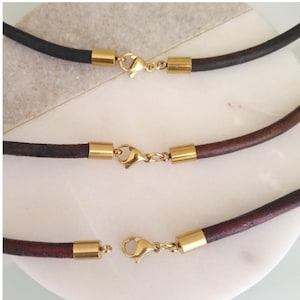 Peut inclure: Gros plan sur trois colliers en cuir avec des fermoirs dorés. Les colliers sont de différentes nuances de marron et de noir. Les fermoirs sont reliés au cuir par des tubes métalliques dorés.
