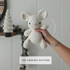 Puede incluir: Un conejito blanco de ganchillo con orejas grandes, ojos negros y nariz rosa se sostiene frente a una pared blanca. El texto "PDF CROCHET PATTERN" se muestra en un rectángulo blanco debajo del conejito. El logotipo de The Cozy Stitch es visible.