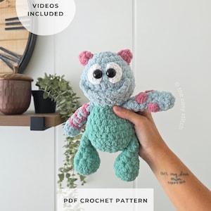 May include: A plush monster toy made of light blue and teal yarn with pink spots on the arms and ears. The creature has large black safety eyes with white felt backing. The image includes the text HELPFUL VIDEOS INCLUDED and PDF CROCHET PATTERN.