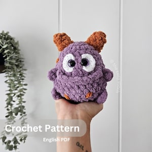 Puede incluir: Un monstruo de ganchillo morado hecho a mano con detalles naranjas y dos cuernos naranjas. El monstruo tiene grandes ojos con bordes blancos. El texto "Crochet Pattern" y "English PDF" son visibles. El monstruo está en una mano.