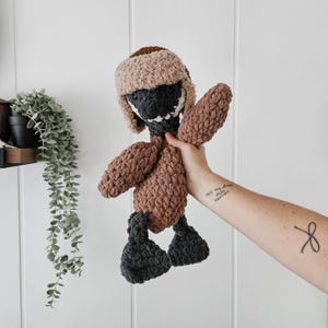 May include: A crocheted stuffed animal in the shape of a duck wearing a brown and white crocheted hat. The duck is brown with dark gray feet and a black beak.