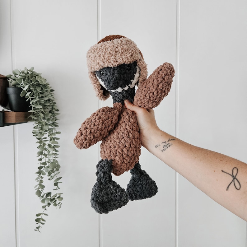 May include: A crocheted stuffed animal in the shape of a duck wearing a brown and white crocheted hat. The duck is brown with dark gray feet and a black beak.