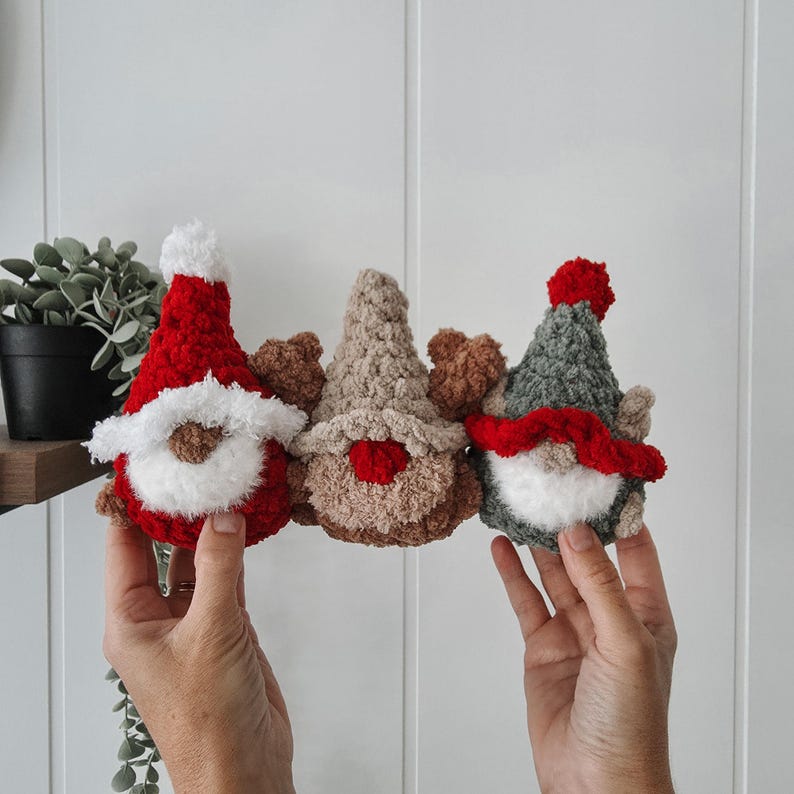 Pode incluir: Tr&ecirc;s gnomos de croch&ecirc; feitos &agrave; m&atilde;o em vermelho, bege e cinza. Cada gnomo tem uma barba fofa, um chap&eacute;u pontudo e pequenos chifres. Os gnomos s&atilde;o apresentados em um fundo branco, sugerindo um tema decorativo ou festivo.