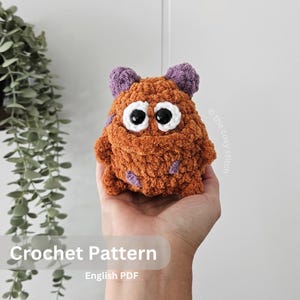 May include: A handmade orange crochet monster with purple accents and large white-rimmed eyes. The plush toy has a textured, fuzzy appearance and two small purple ears. The text "Crochet Pattern" and "English PDF" are visible in the image.
