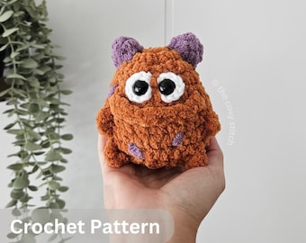 Pode incluir: Um monstro de crochê laranja feito à mão com detalhes roxos e olhos grandes com bordas brancas. O brinquedo de pelúcia tem uma aparência texturizada e difusa e duas pequenas orelhas roxas. O texto "Crochet Pattern" e "English PDF" são visíveis.