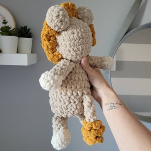 May include: A crocheted lion plush toy with a beige body and yellow mane. The toy is being held by a hand with a tattoo.