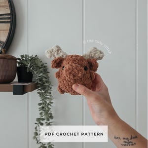 May include: A small, brown crocheted moose with beige antlers is held up in front of a white wall. The moose has small black eyes. The text "PDF CROCHET PATTERN" is displayed in a white rectangle below the moose.
