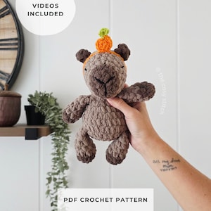 May include: A brown crocheted plush animal toy featuring a small orange pumpkin with a green stem attached to its head. The toy has a soft, textured yarn appearance. Text on the image reads HELPFUL VIDEOS INCLUDED and PDF CROCHET PATTERN.