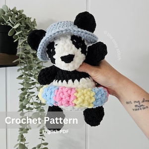 Può includere: Un orsetto panda all'uncinetto fatto a mano, con pelliccia bianca e nera, un cappello azzurro e una fascia colorata. Il testo "Crochet Pattern" e "English PDF" sono visibili. Un giocattolo artigianale.