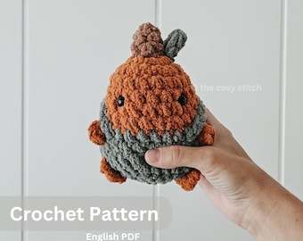 May include: Crochet pattern for a small, orange and gray amigurumi toy. The toy has a round body with a small, brown pom-pom on top. The pattern is available as an English PDF download. "The cosy stitch" is written on the image.